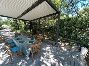 An outdoor dining area with a covered table and chairs, surrounded by greenery and stone walls. - Spacious Provencal villa in absolute calm. (Roquefort-les-Pins)
