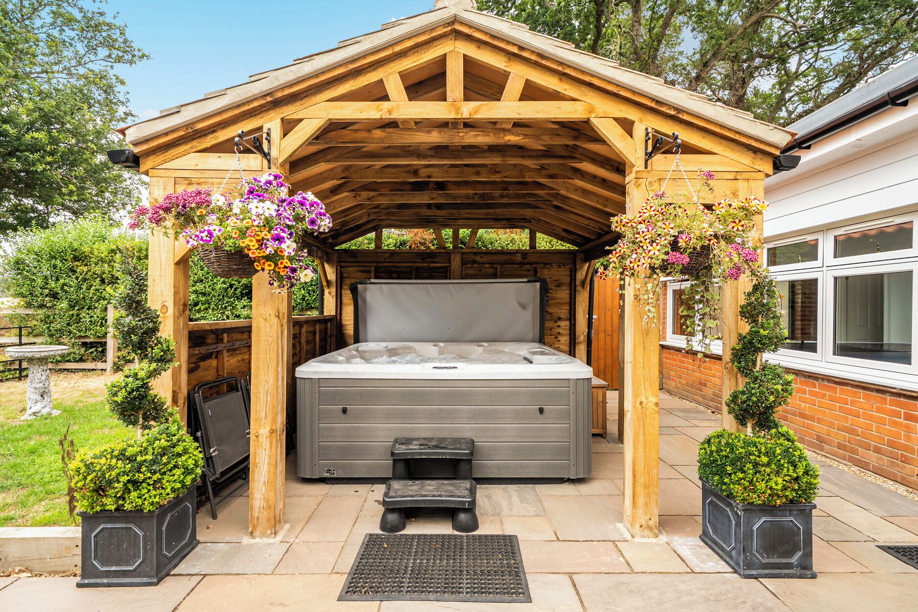 Un bain à remous installé sous une pergola en bois, agrémentée de suspensions fleuries et entouré de plantes en pot.