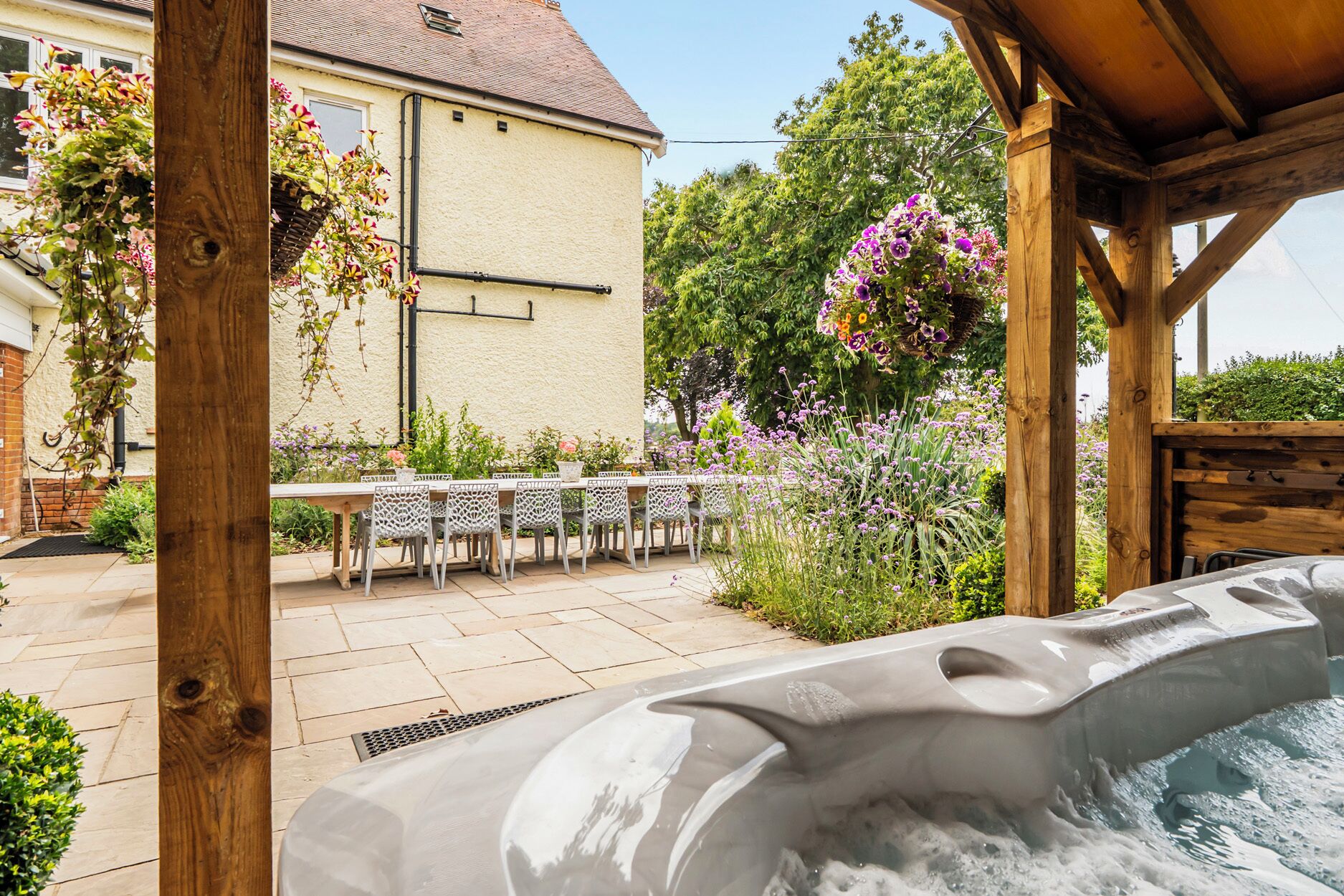 A hot tub in a covered outdoor area with a wooden pergola, a table with chairs, and a building in the background.