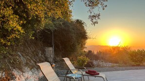 Une vue sur le coucher de soleil, avec des chaises longues et une table, le tout entouré d’un paysage naturel et d’un mur de pierre.