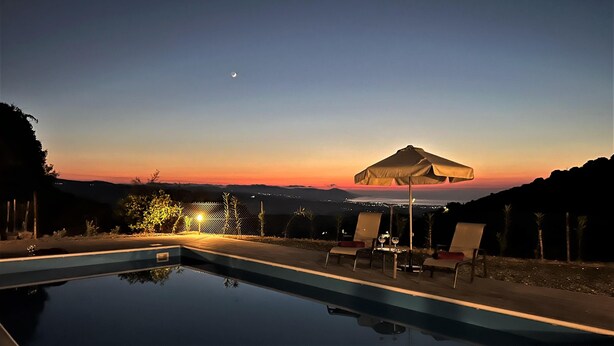 Une piscine entourée d’une clôture et d’arbres, agrémentée de chaises longues et d’un parasol, avec une vue sur le coucher de soleil en arrière-plan.