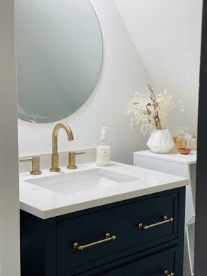 Een badkamer met een witte wastafel, gouden kraanwerk, een grote spiegel, een donkerblauwe kast met gouden knoppen, en een vaas met gedroogde bloemen.