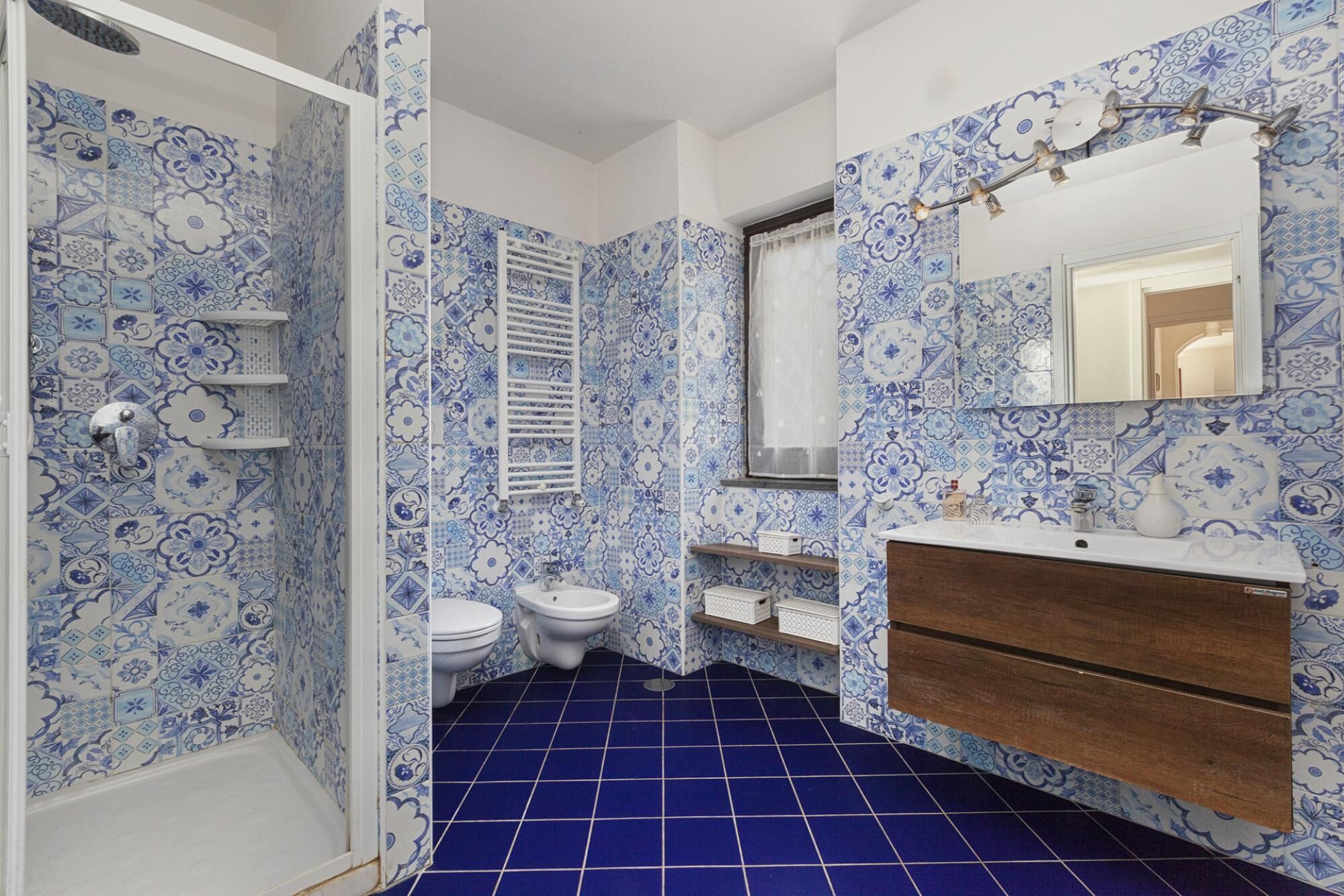 A bathroom with blue and white patterned tiles, a white bathtub, two toilets, a wooden vanity with a mirror, and a blue tiled floor.