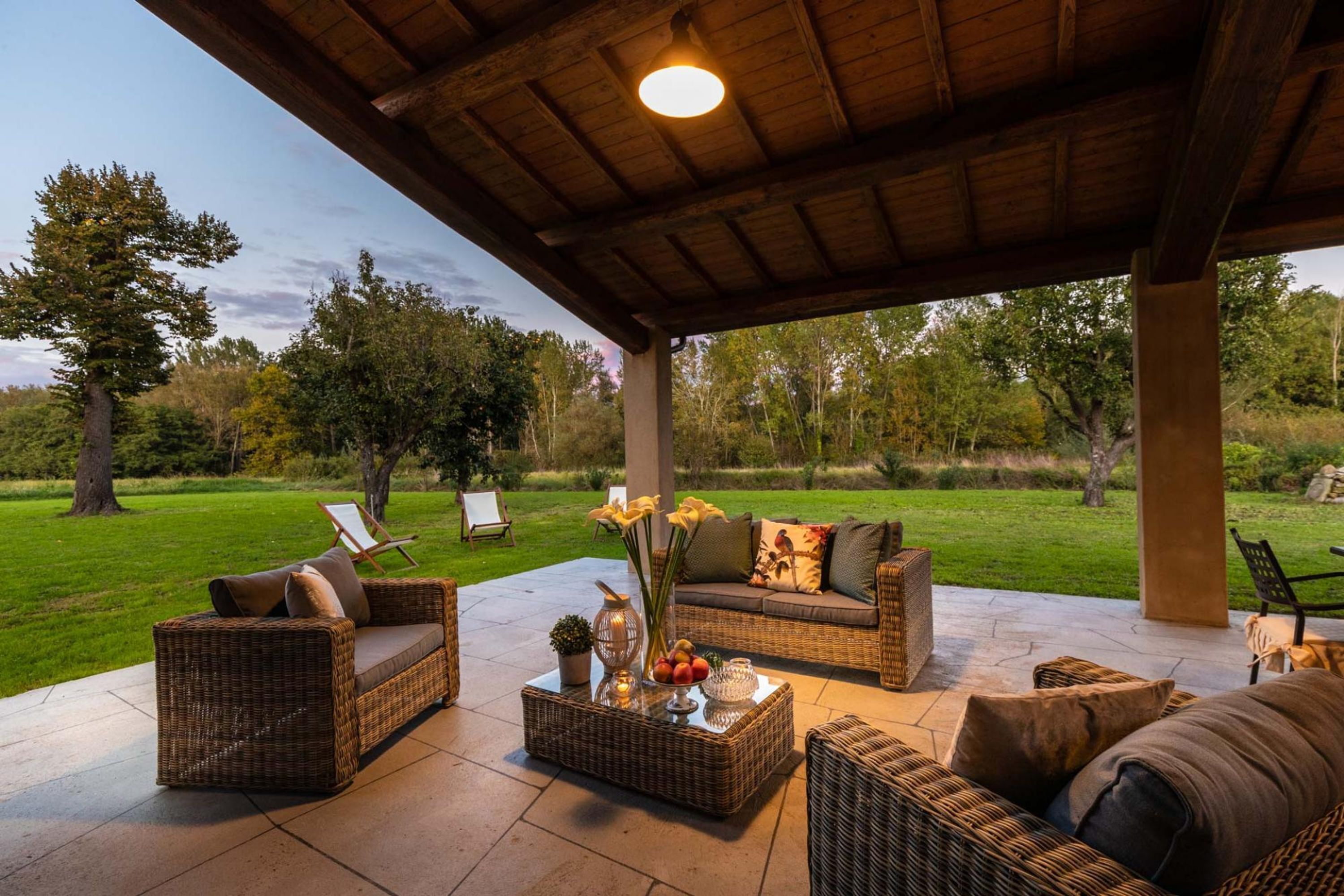 An outdoor patio area with wicker furniture, a glass coffee table, and a view of a grassy landscape.