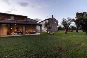 Un edificio rústico de piedra con techo de tejas, un patio con mobiliario exterior y un jardín cubierto de césped.
