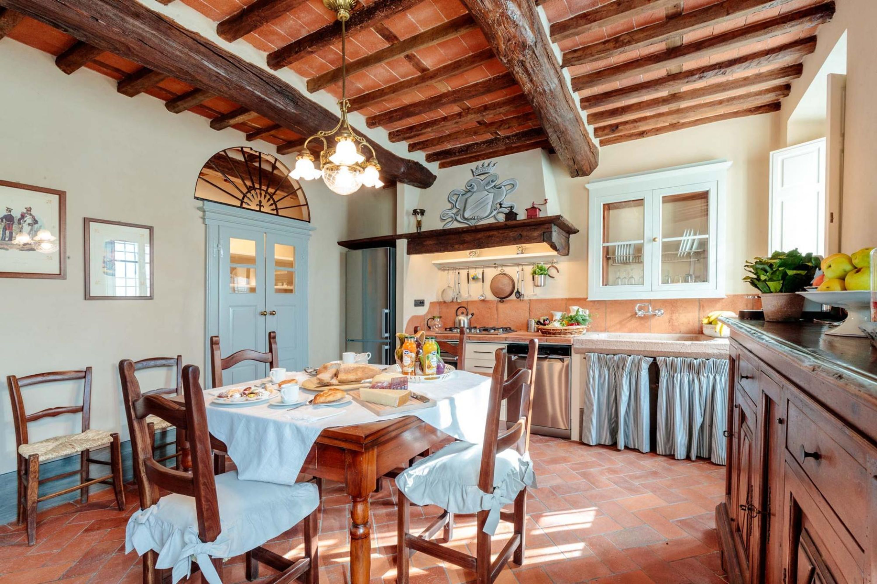 Una cucina rustica con travi in legno, un tavolo da pranzo apparecchiato con piatti e pane, e una rastrelliera per pentole sospesa sopra il fornello.