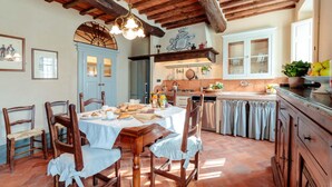 Cozinha rústica com vigas de madeira, mesa de jantar posta com pratos e pão, e um suporte para panelas pendurado acima do fogão.