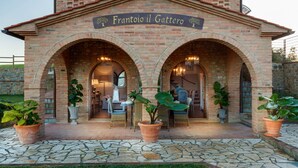 Un bâtiment en briques doté de deux entrées en arc de cercle, de plantes en pot et d’une enseigne indiquant « Frantoio il Gattero ».