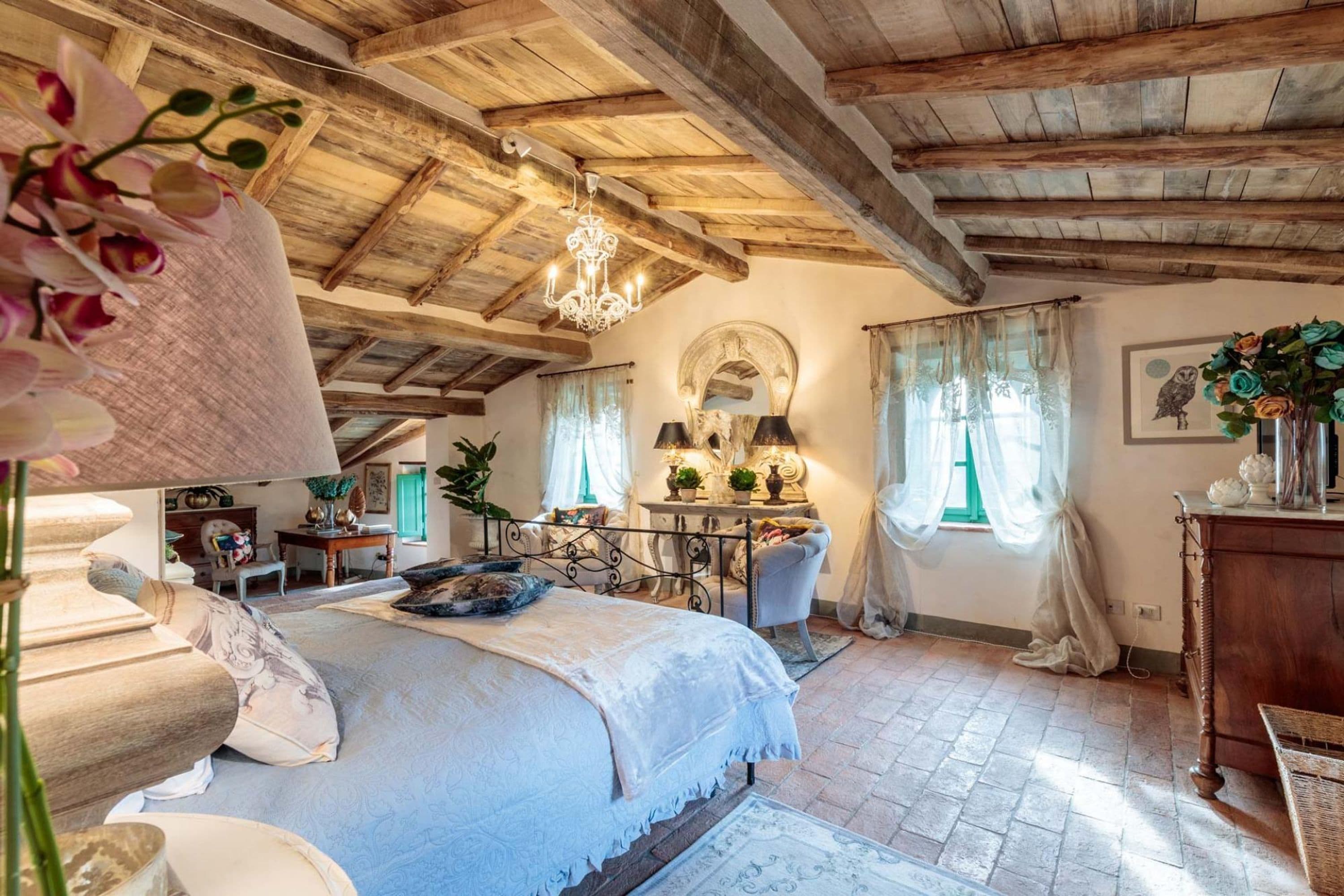 A rustic bedroom with exposed wooden beams, a chandelier, a bed with a floral pillow, a desk with a mirror, and a vase of flowers.