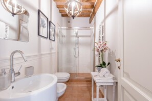 A bathroom with a large white sink, a toilet, a glass-enclosed shower, and a small white vanity with a vase of flowers. - Villa Hilary in Lucca (Lucca)