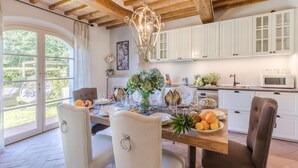 A dining area with a wooden table set for four, white chairs, a chandelier, and a view of the outdoors through a glass door. - Villa Hilary in Lucca (Lucca)