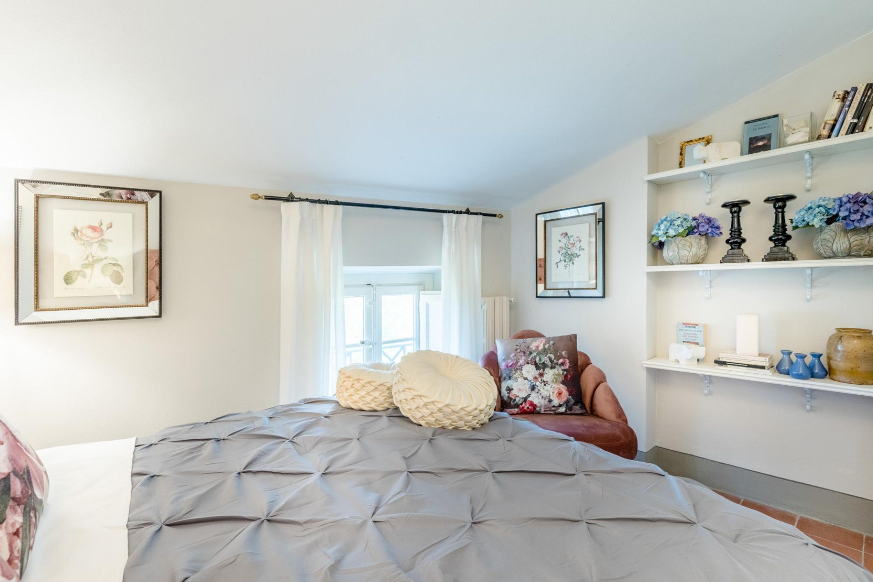 A bedroom with a bed, pillows, a chair, a window with curtains, and shelves with decorative items.