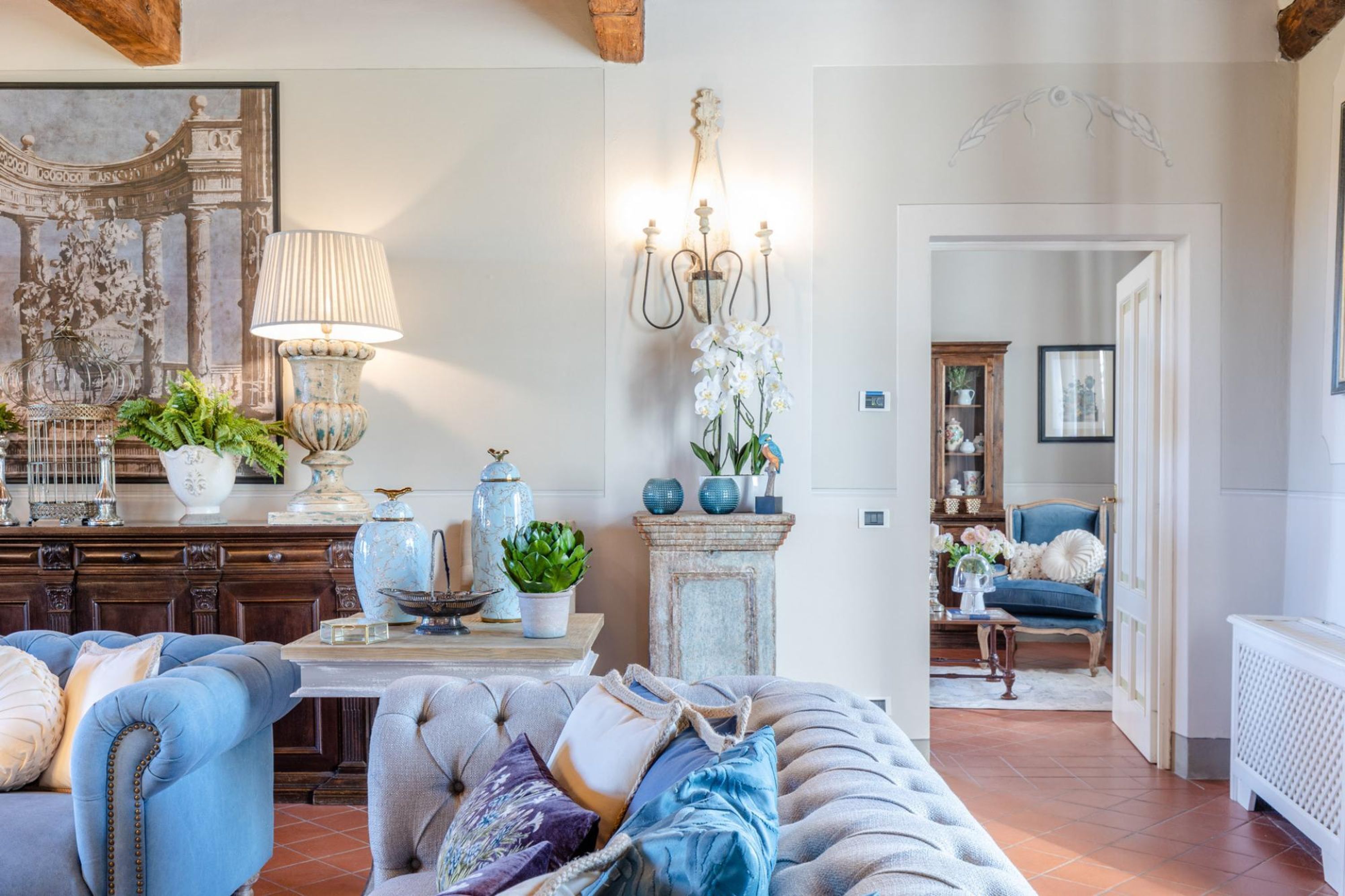 Uma sala de estar com um sofá azul, uma poltrona estofada, uma mesa com objetos decorativos, uma luminária de parede e um lustre.
