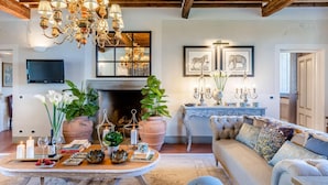 A cozy living room with a fireplace, a large sofa, and a coffee table. There are potted plants, candles, and a chandelier. - Fattoria Camigliano Wine Estate (Capannori)