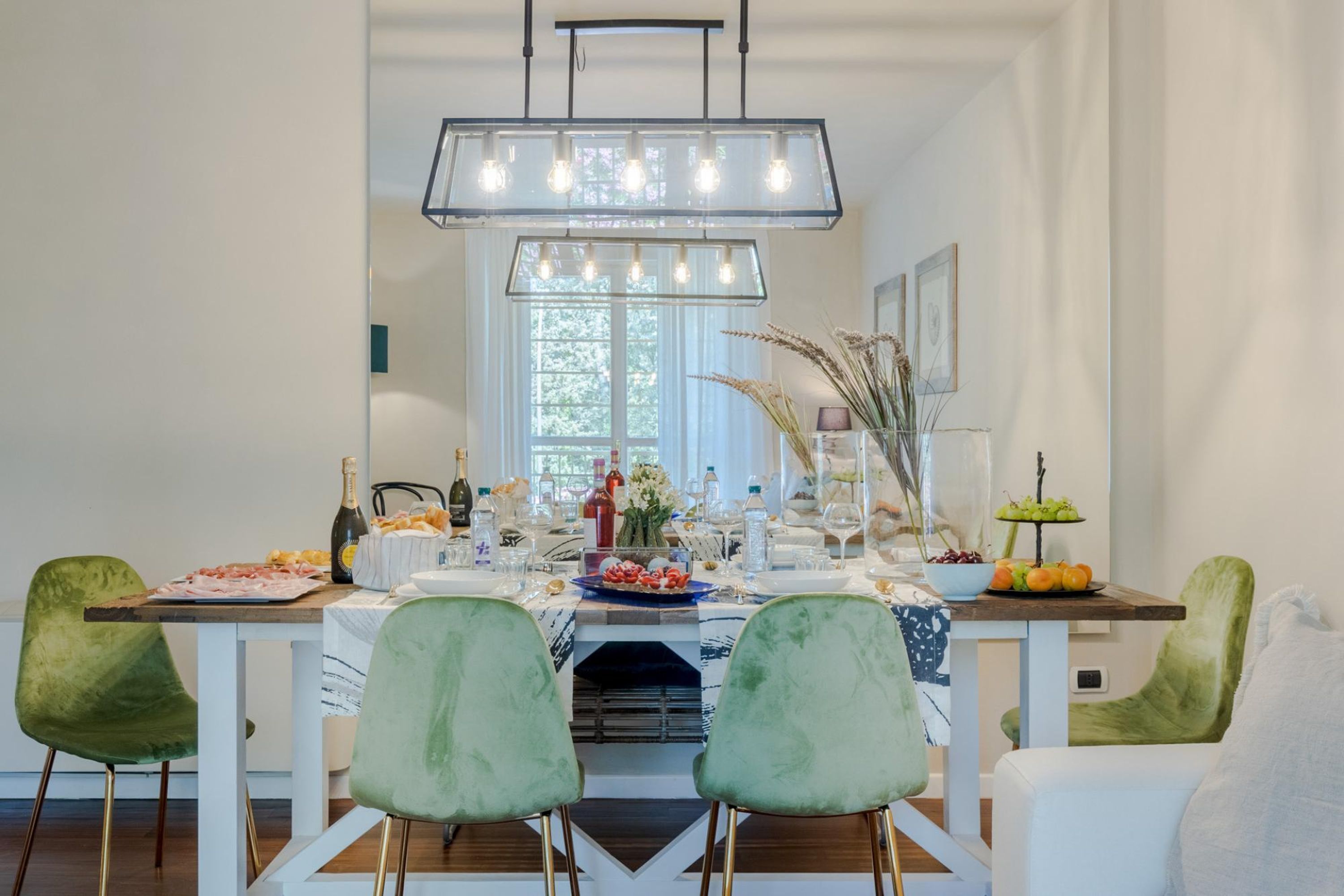 A dining area with a long table set for a meal, surrounded by green chairs, and a modern chandelier above.