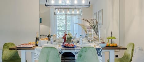 A dining area with a long table set for a meal, surrounded by green chairs, and a modern chandelier above.
