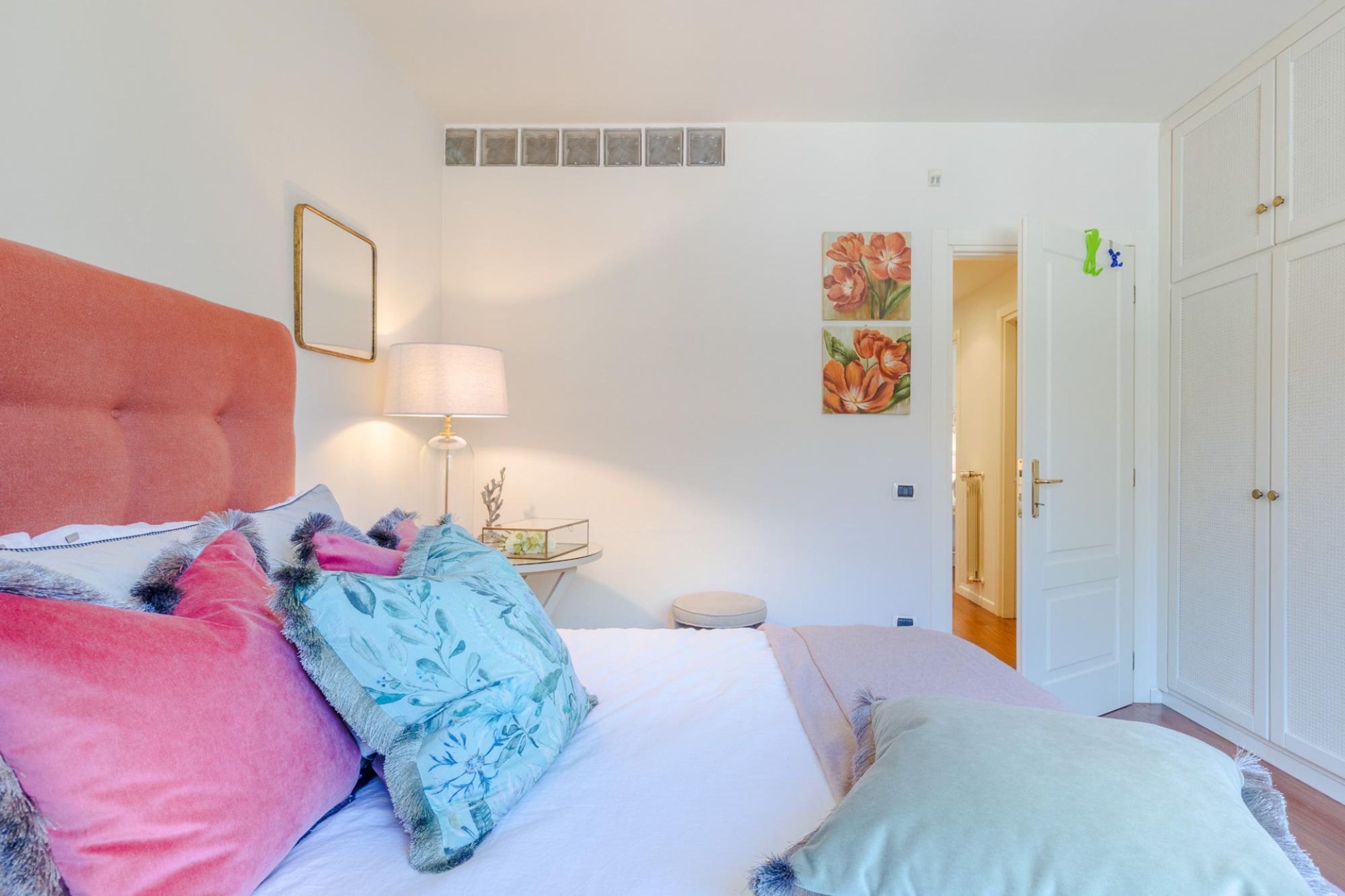 A bedroom with a bed, pillows, a bedside table with a lamp, a small round stool, and a wardrobe.