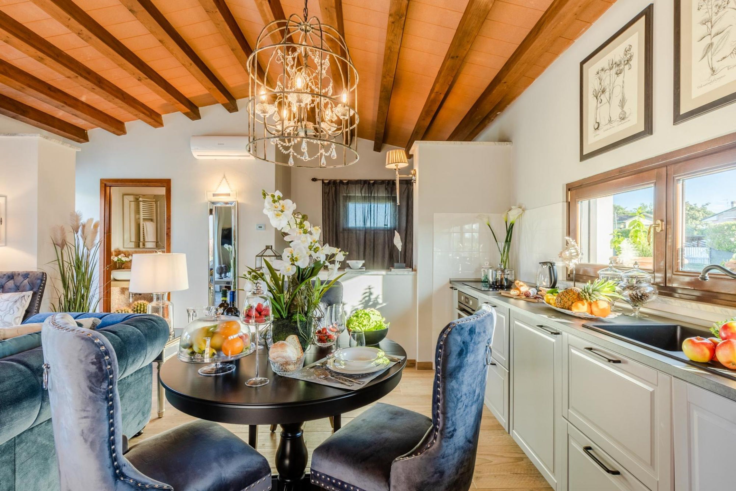 Un coin repas avec une table ronde, deux chaises bleues à revêtement rembourré, un lustre et une cuisine équipée d’un évier et d’un présentoir à fruits.
