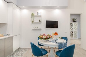 A modern kitchen with white cabinetry, a dining area with a round table and blue chairs, a flat-screen TV mounted on the wall, and a small plant on a shelf.