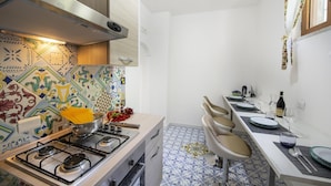 A compact kitchen with a gas stove, a sink, and a dining area with a table set for two. - Amore Rentals - Casa Barbera A (Positano)
