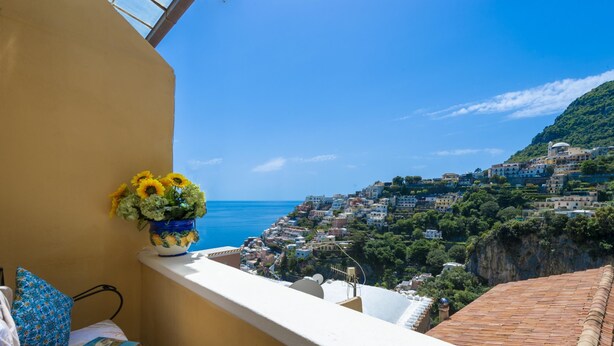 A balcony with a view of a coastal town, a vase with sunflowers, and a patterned cushion. - Amore Rentals - Casa Barbera A (Positano)