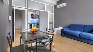 Egy modern nappali, melyben egy üveg asztallapos étkezőasztal, egy kék kanapé és egy nyitott ajtón keresztül belátható konyha található.