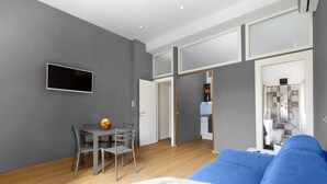 Egy modern nappali, szürke színű kanapéval, étkezőasztallal és székekkel, valamint egy nyitott ajtón keresztül belátható konyhasarokkal.