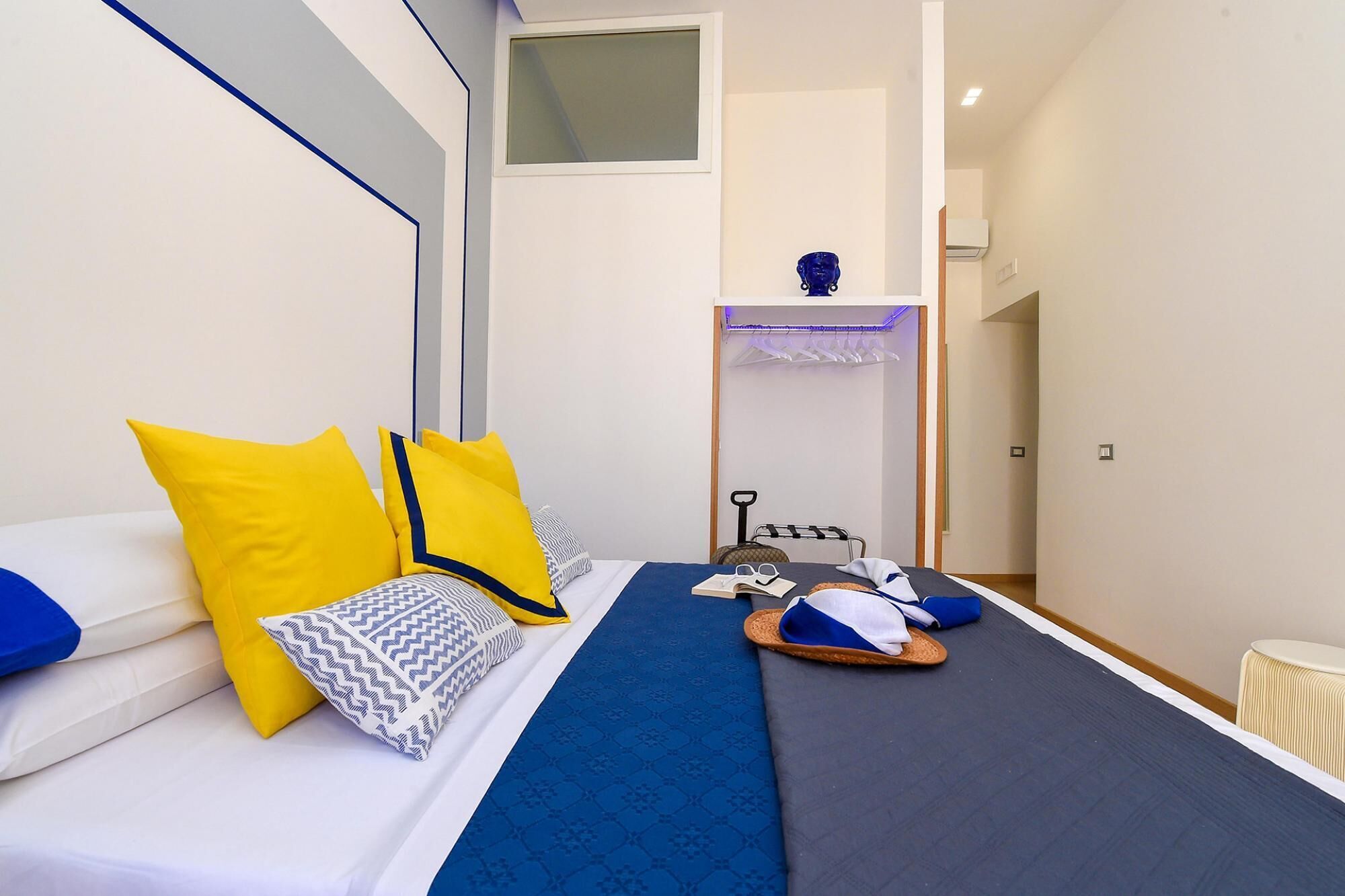 A bedroom with a bed, pillows, a blue blanket, and a small table with a hat and sunglasses.