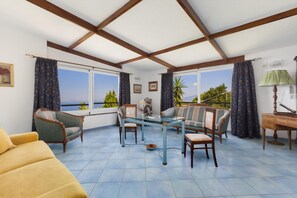 A sunlit room with a view of the sea, featuring a sofa, chairs, a table, and a lamp.