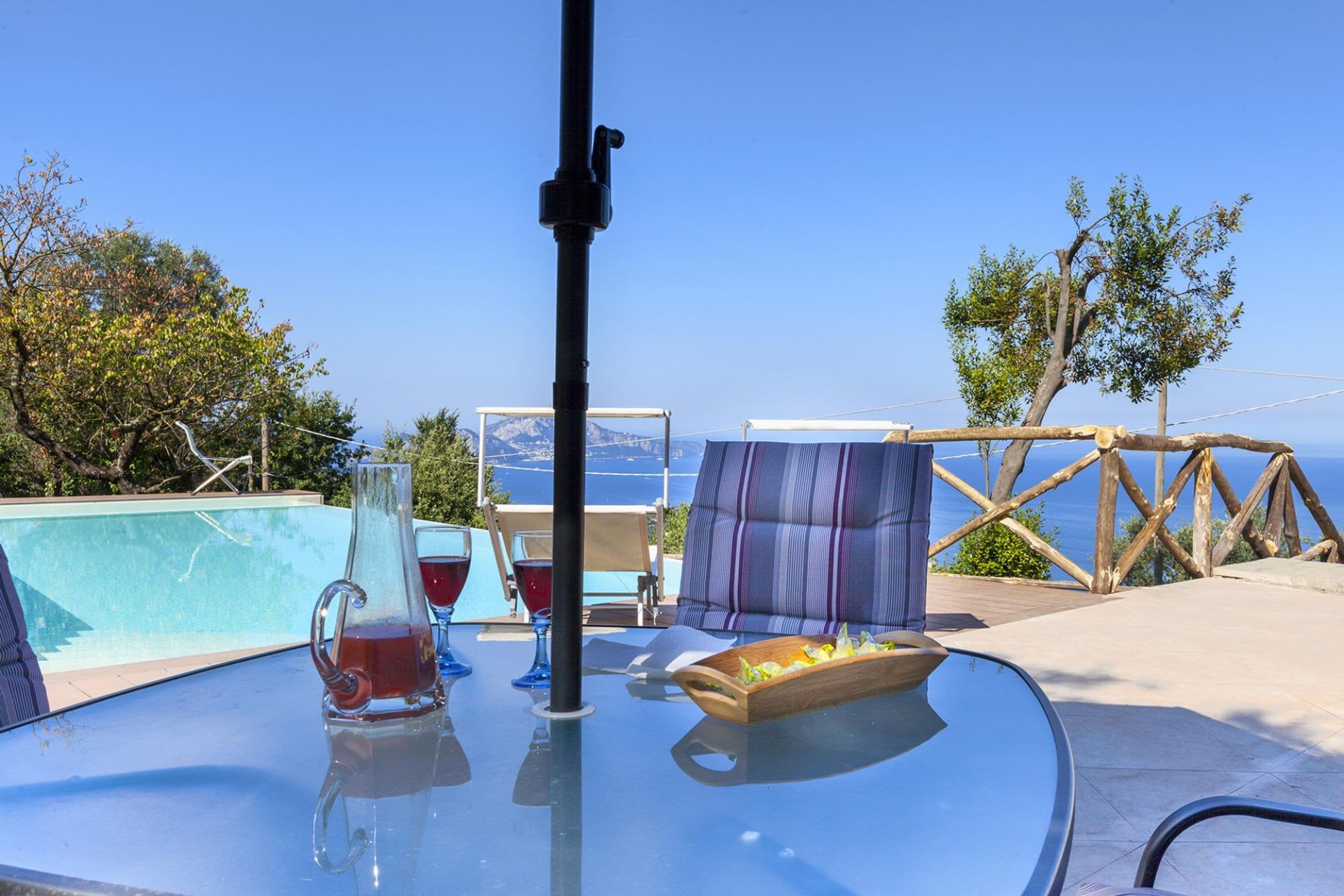 A glass table with a pitcher of red drink, two glasses, and a wooden tray with food, overlooking a pool and the sea.