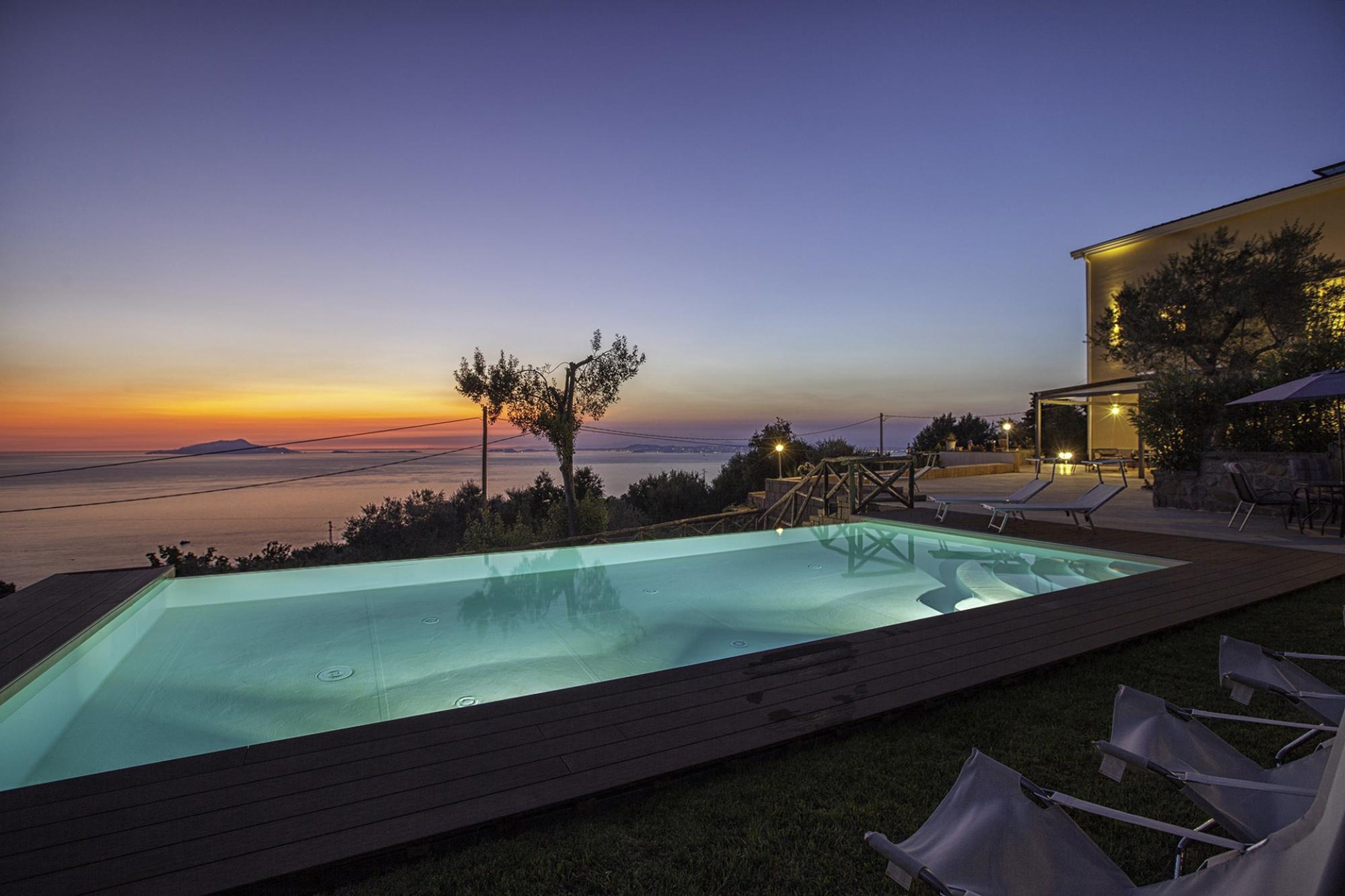 Une piscine avec vue sur la mer au coucher du soleil, entourée de chaises longues et d’un bâtiment doté d’un éclairage extérieur.
