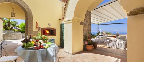 Un patio con mesa lista para una comida, un horno de ladrillo y vista al mar.