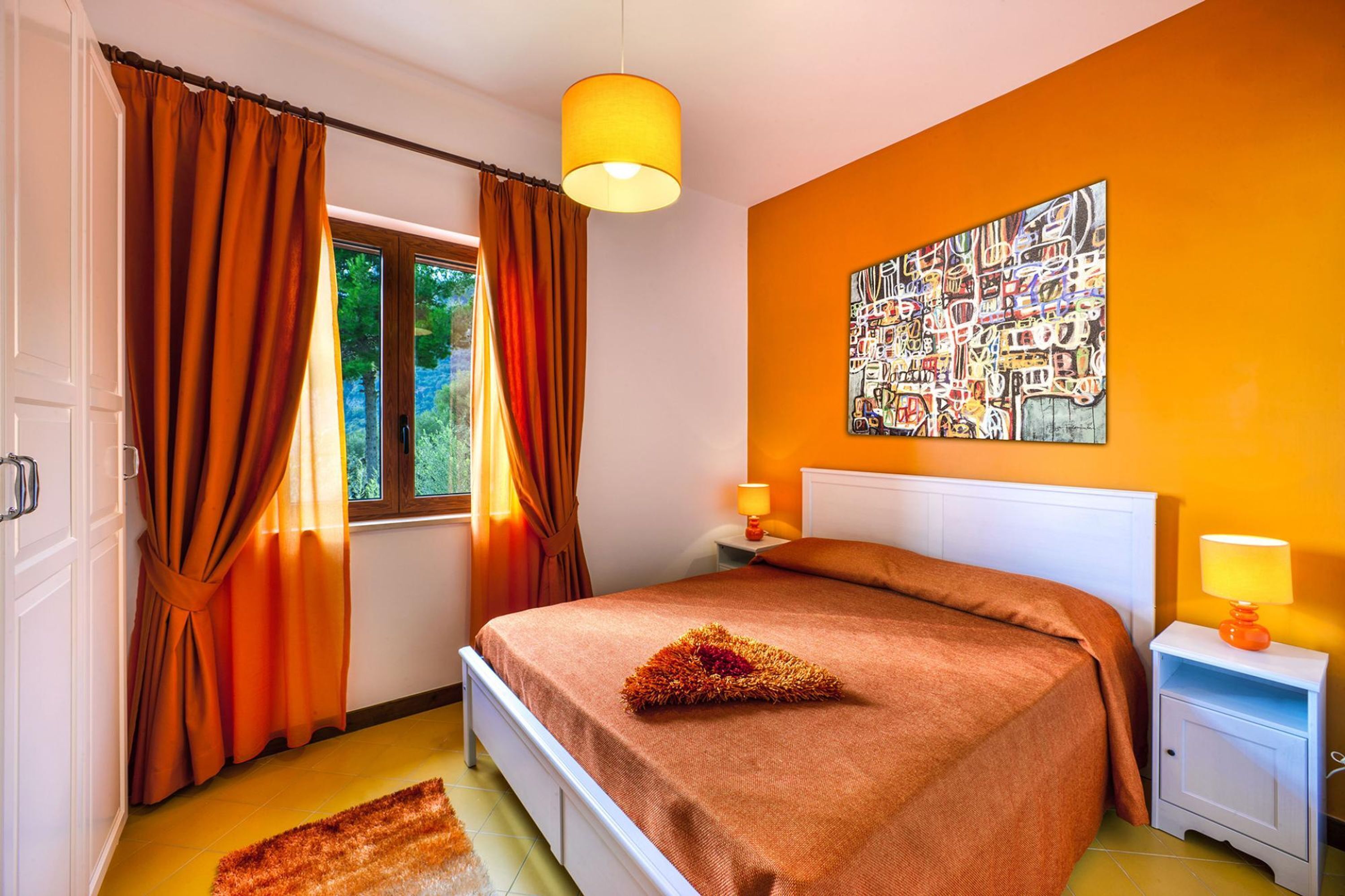 Un dormitorio con cama, cortinas, mesita de noche, lámpara y un cuadro en la pared.