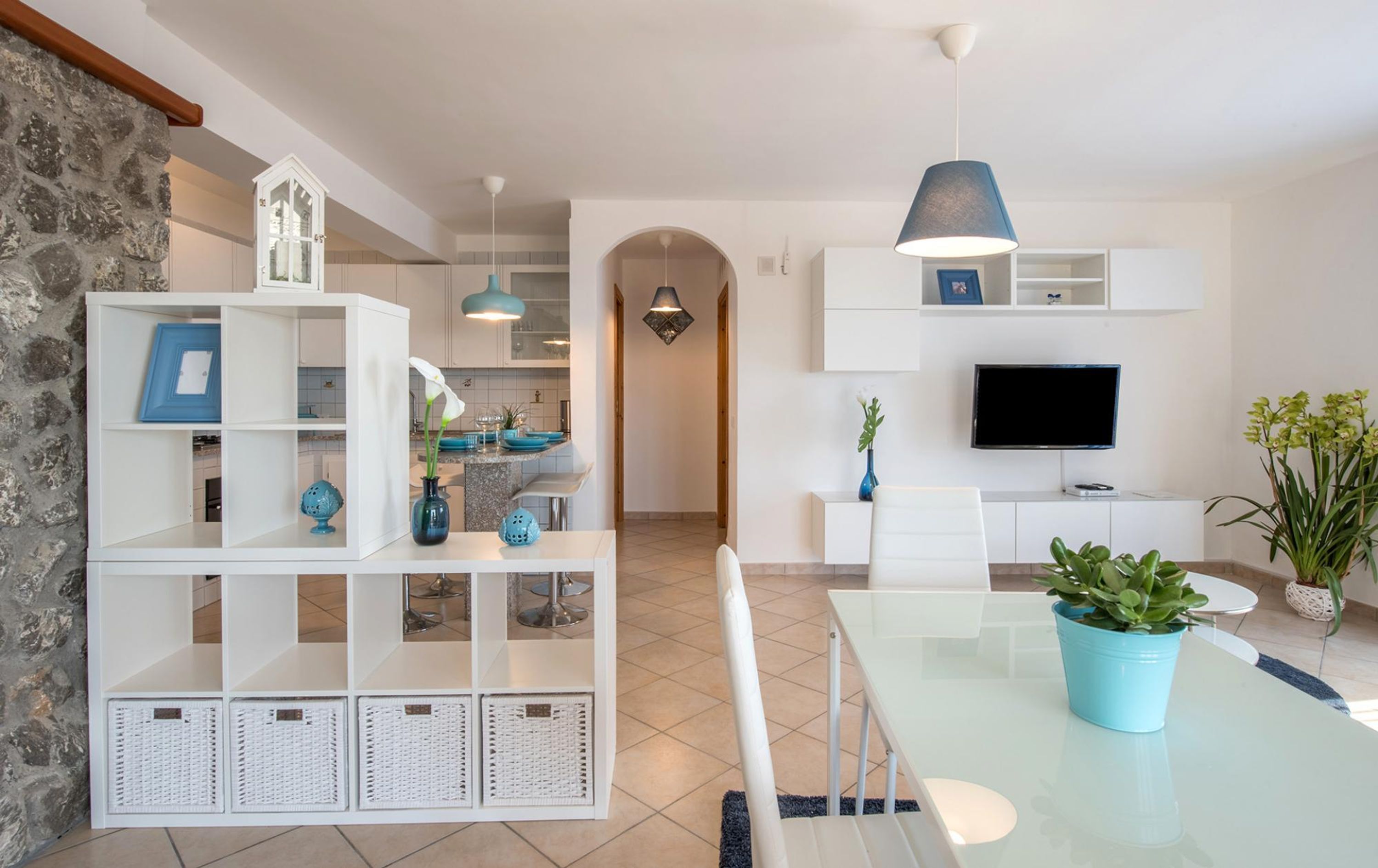 Een moderne woonkamer met een glazen eettafel, witte meubels, een televisie en decoratieve planten.