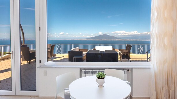A balcony with a table and chairs, a potted plant, and a view of the sea and a mountain. - Villa Ado in Sorrento (Sorrento)
