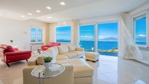 A modern living room with a beige sofa, a red armchair, and a glass coffee table. Large windows offer a view of the sea and a distant mountain. - Villa Ado in Sorrento (Sorrento)