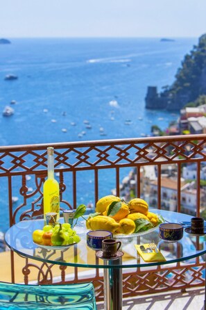 Uma mesa de vidro com limões, uma garrafa e uma taça de uvas numa varanda com vista para o mar.