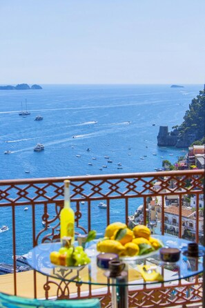 Varanda com mesa posta para refeições, vista para o mar costeiro, barcos e um penhasco rochoso.