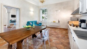 Un vaste espace de vie comprenant une table à manger en bois, des chaises en plastique transparent, un canapé bleu et une cuisine équipée d’appareils électroménagers modernes.