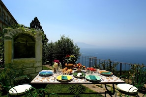 Une table à manger installée à l’extérieur, avec des assiettes, des verres et un saladier rempli de fruits, offrant une vue panoramique sur la mer.