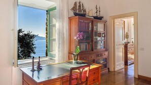 A room with a large window overlooking a body of water, a wooden cabinet with books, and a table with a vase of flowers. - Villa Alma in Massa Lubrense (Massa Lubrense)
