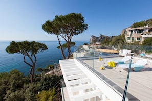 Une terrasse sur le toit offrant une vue dégagée sur la mer, un espace piscine avec des chaises longues et, au loin, un bâtiment en pierre.