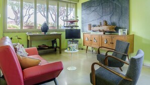 A living room with a red sofa, grey armchairs, a wooden cabinet, a television, and a large window with a view of trees. - La Casa Matta in Viareggio (Viareggio)