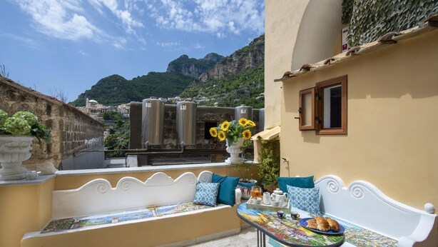 A terrace with a white bench and table, a view of mountains, and a building with a tiled roof. - Amore Rentals - Casa Barbera B (Positano)