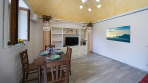 A dining area with a wooden table set for four, a television on a white cabinet, and a painting of a seascape on the wall. - Amore Rentals - Casa Barbera B (Positano)