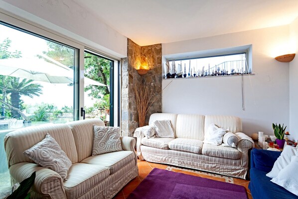 A living room with two sofas, a stone accent wall, a window with a view of greenery, and a shelf with decorative items. - Villa The Stone on the Sea (Massa Lubrense)