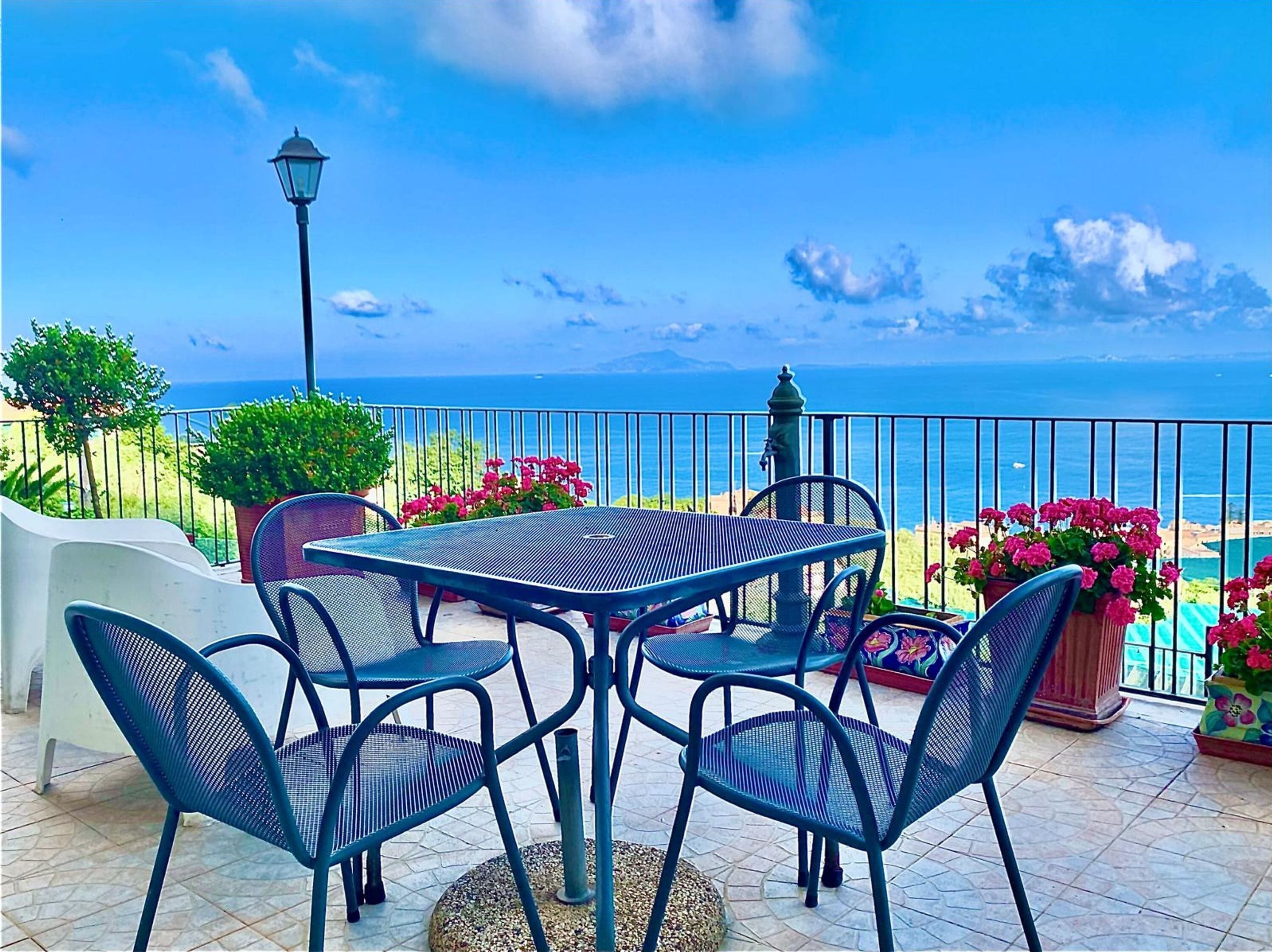 A terrace with a table and chairs, a lamp, and potted plants with flowers, overlooking a coastal view.