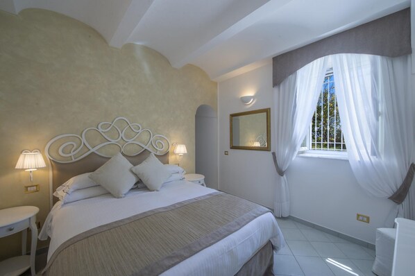 Un dormitorio con cama, dos mesitas de noche, un tocador, una ventana con cortinas, un espejo en la pared y una lámpara.