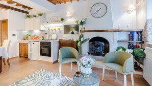 Una sala de estar acogedora con chimenea, dos sillones, un comedor con mesa y sillas, y una cocina con armarios blancos y salpicadero de azulejos.