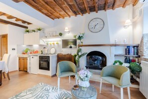 Ein gemütliches Wohnzimmer mit Kamin, zwei Sesseln, einer Essecke mit Tisch und Stühlen sowie einer Küche mit weißen Schränken und einer gefliesten Spritzschutzwand.